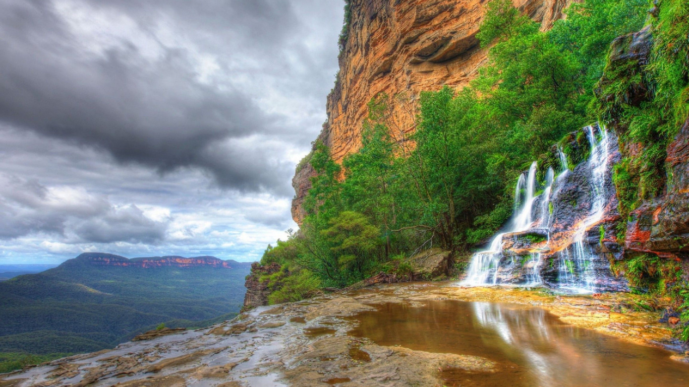 El Agua Cae Entre la Montaña Rocosa Marrón Bajo un Cielo Nublado Gris Durante el Día. Wallpaper in 1366x768 Resolution