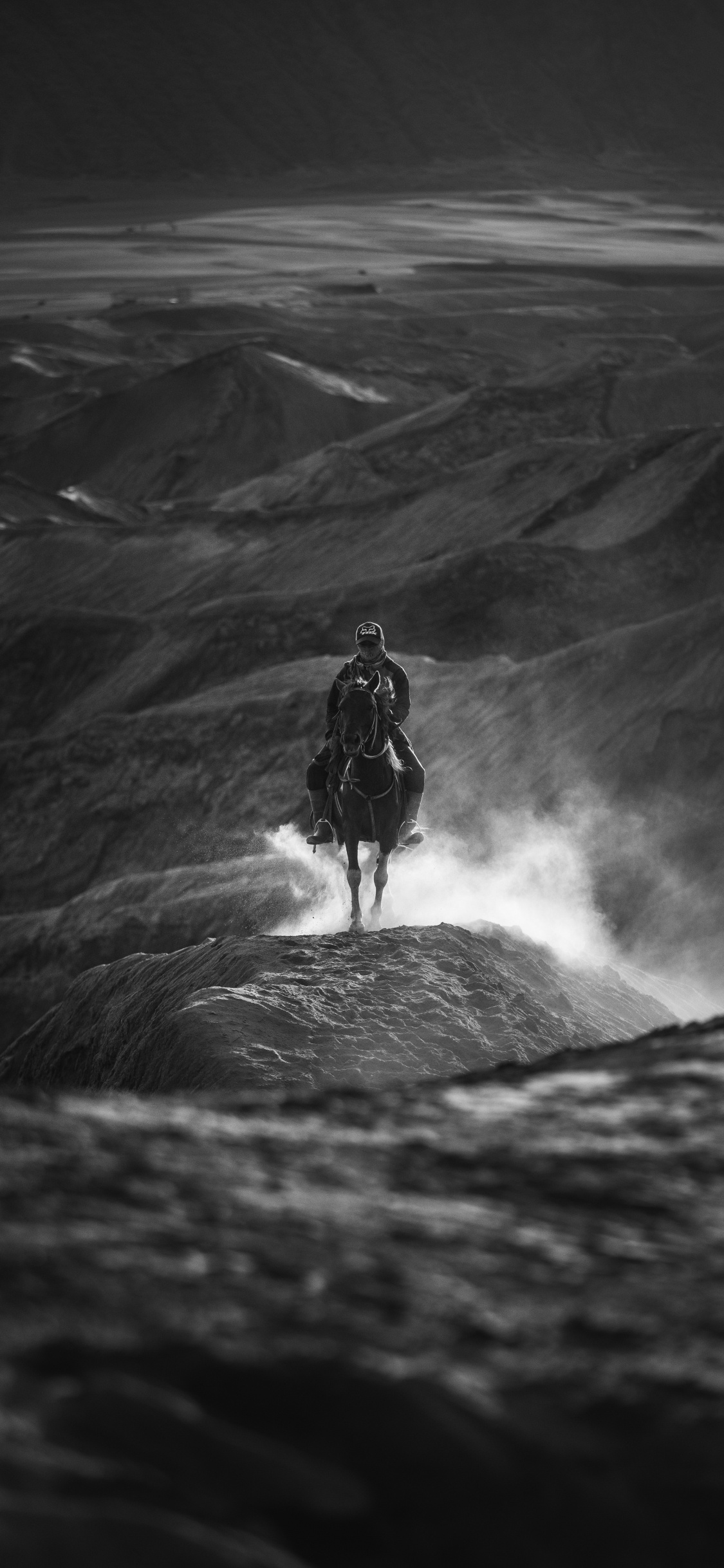 Grayscale Photo of Man in Black Jacket and Pants Standing on Rock Formation. Wallpaper in 1125x2436 Resolution