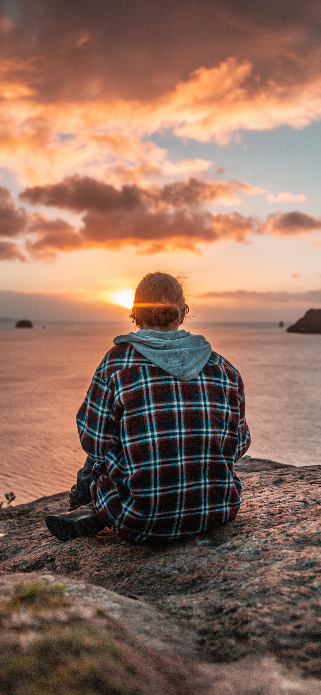 Man in Blue and White Plaid Dress Shirt Sitting on Rock Near Body of Water During. Wallpaper in 1125x2436 Resolution