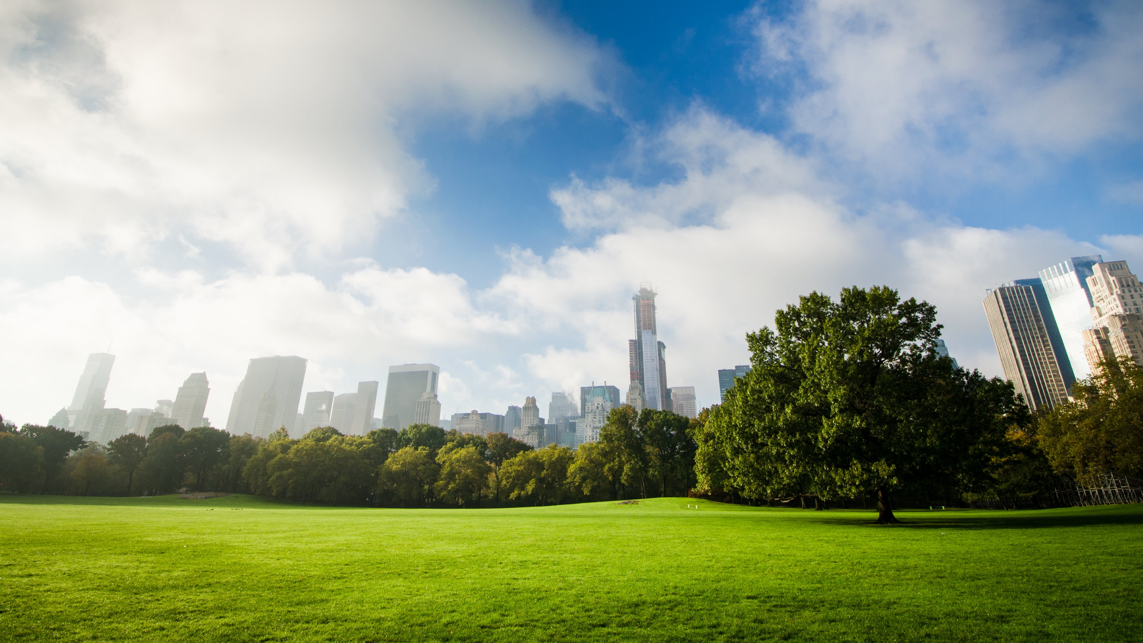 Green Grass Field Near City Buildings Under Blue and White Sunny Cloudy Sky During Daytime. Wallpaper in 3840x2160 Resolution