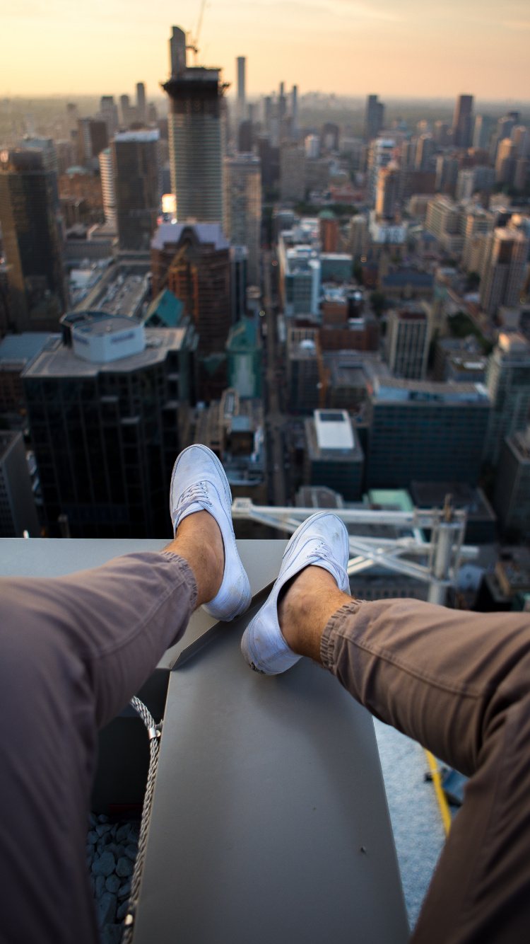 Person in Brown Pants and White Sneakers Sitting on White Building During Daytime. Wallpaper in 750x1334 Resolution