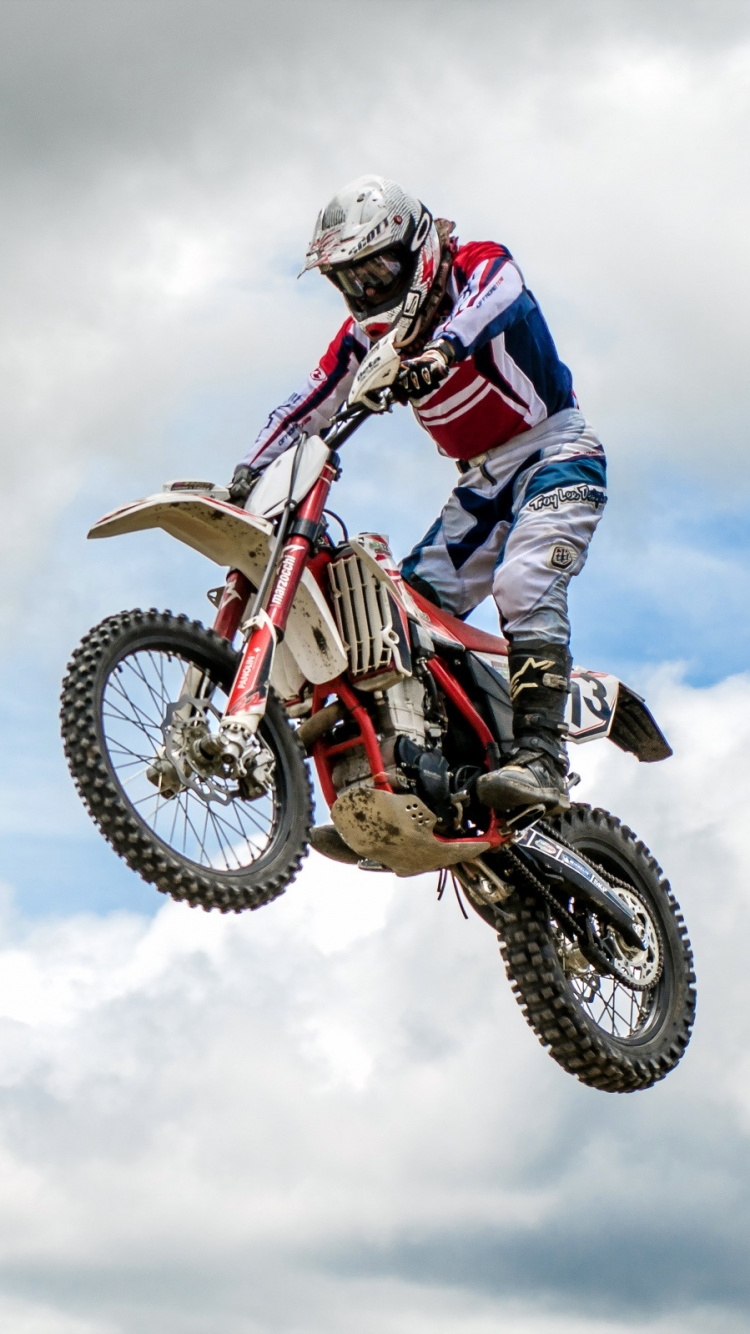 Hombre Montando Motocross Dirt Bike Haciendo Acrobacias Bajo Las Nubes Blancas Durante el Día. Wallpaper in 750x1334 Resolution
