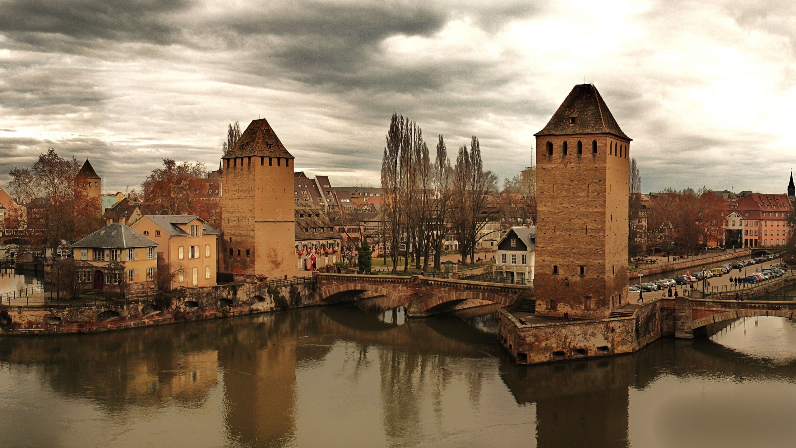 Brown Brick Building Beside River Under Cloudy Sky. Wallpaper in 2560x1440 Resolution