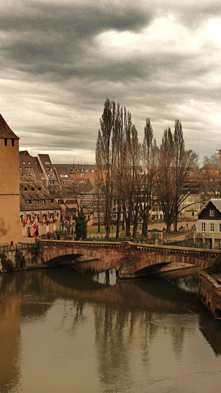 Brown Brick Building Beside River Under Cloudy Sky. Wallpaper in 750x1334 Resolution