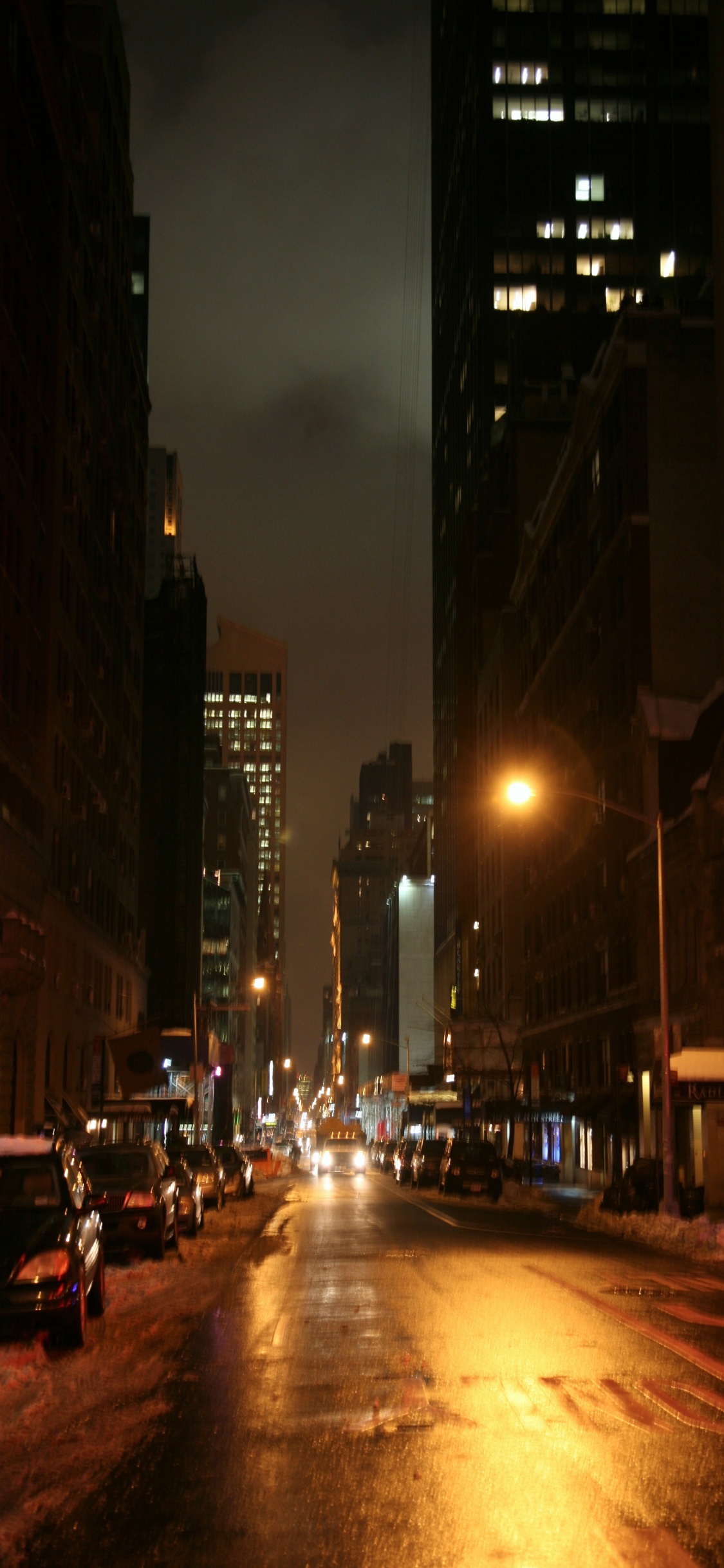 Autos Auf Der Straße Zwischen Hochhäusern Während Der Nacht. Wallpaper in 1125x2436 Resolution