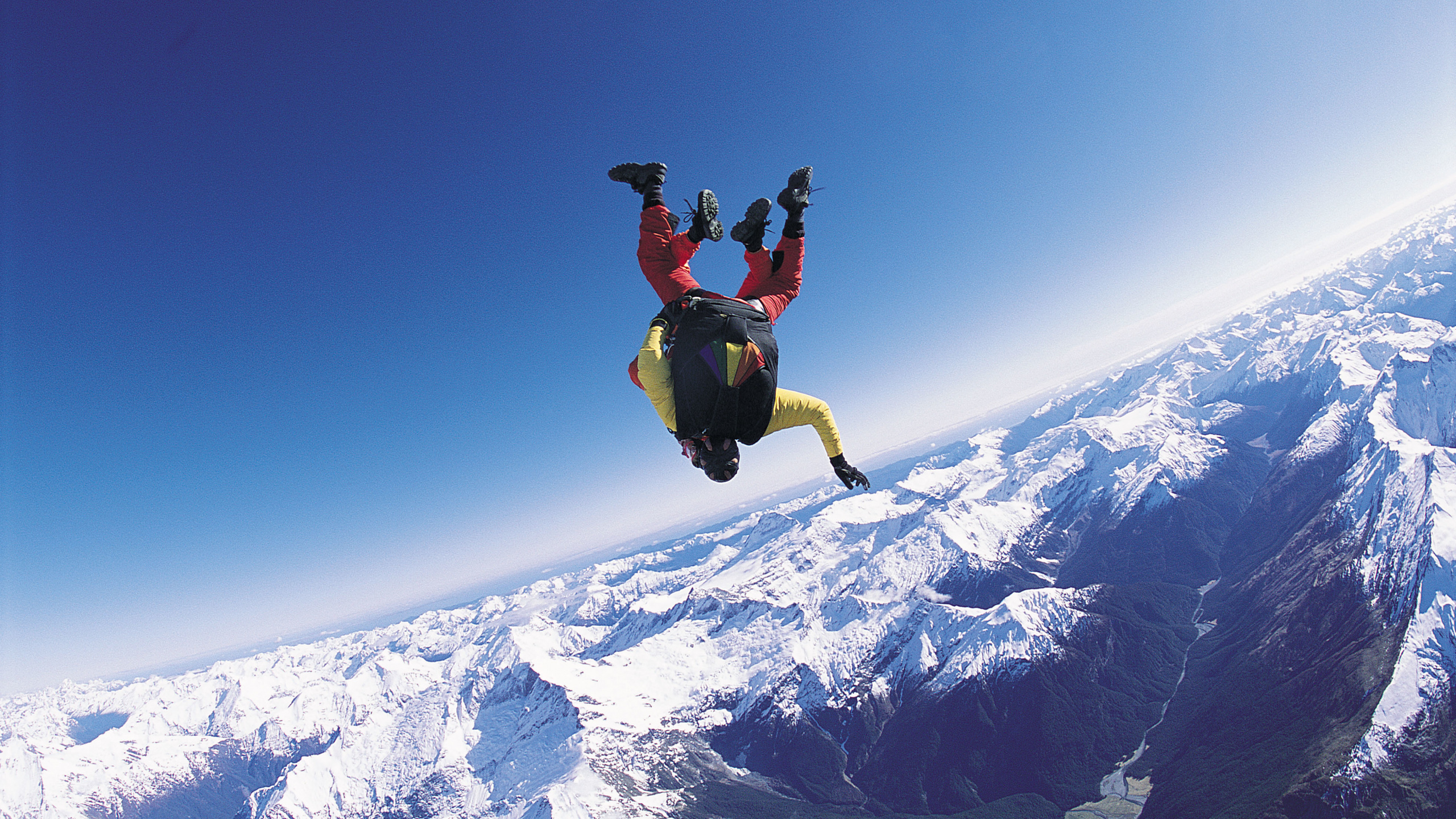 Man in Black Jacket and Black Pants Jumping on Snow Covered Mountain During Daytime. Wallpaper in 2560x1440 Resolution