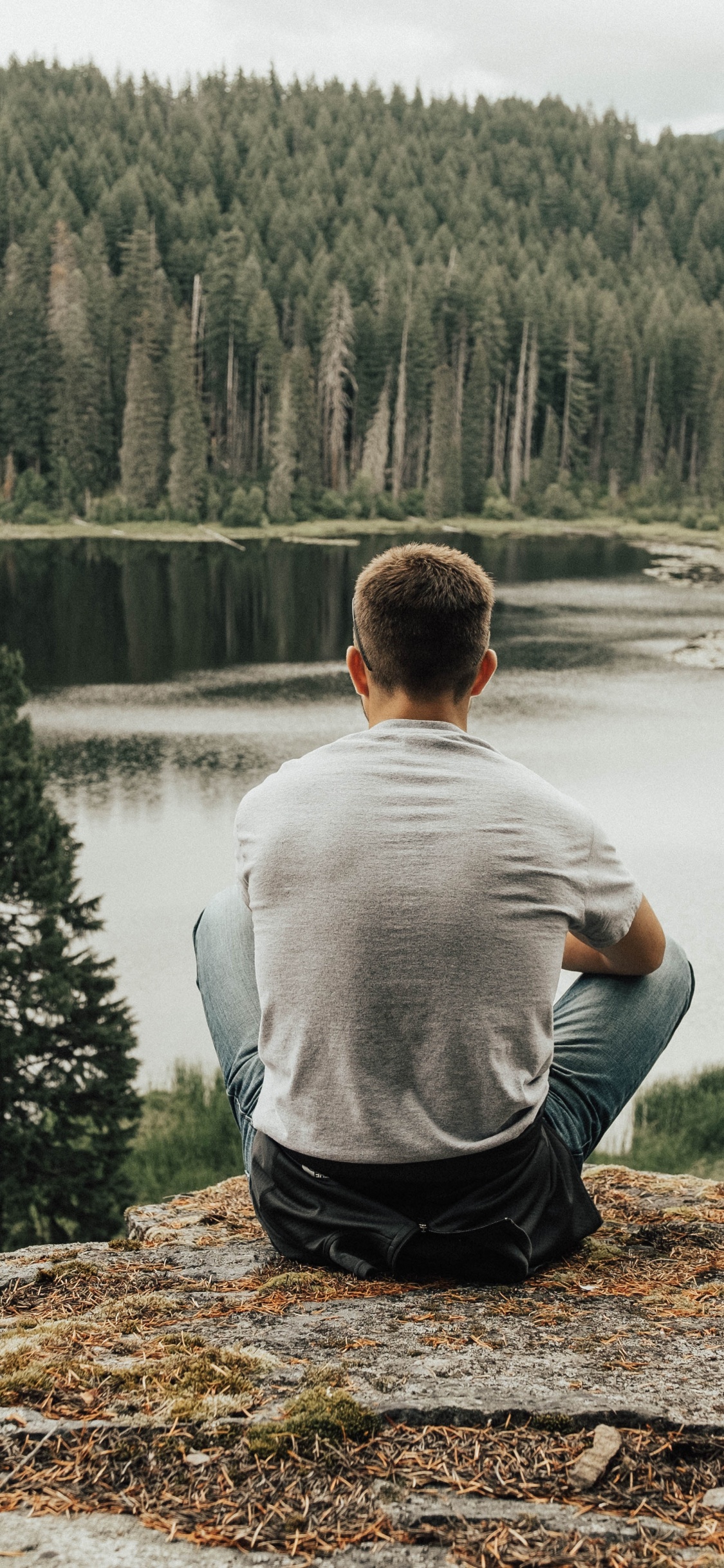 Man in Gray Sweater Sitting on Brown Rock Near Lake During Daytime. Wallpaper in 1125x2436 Resolution