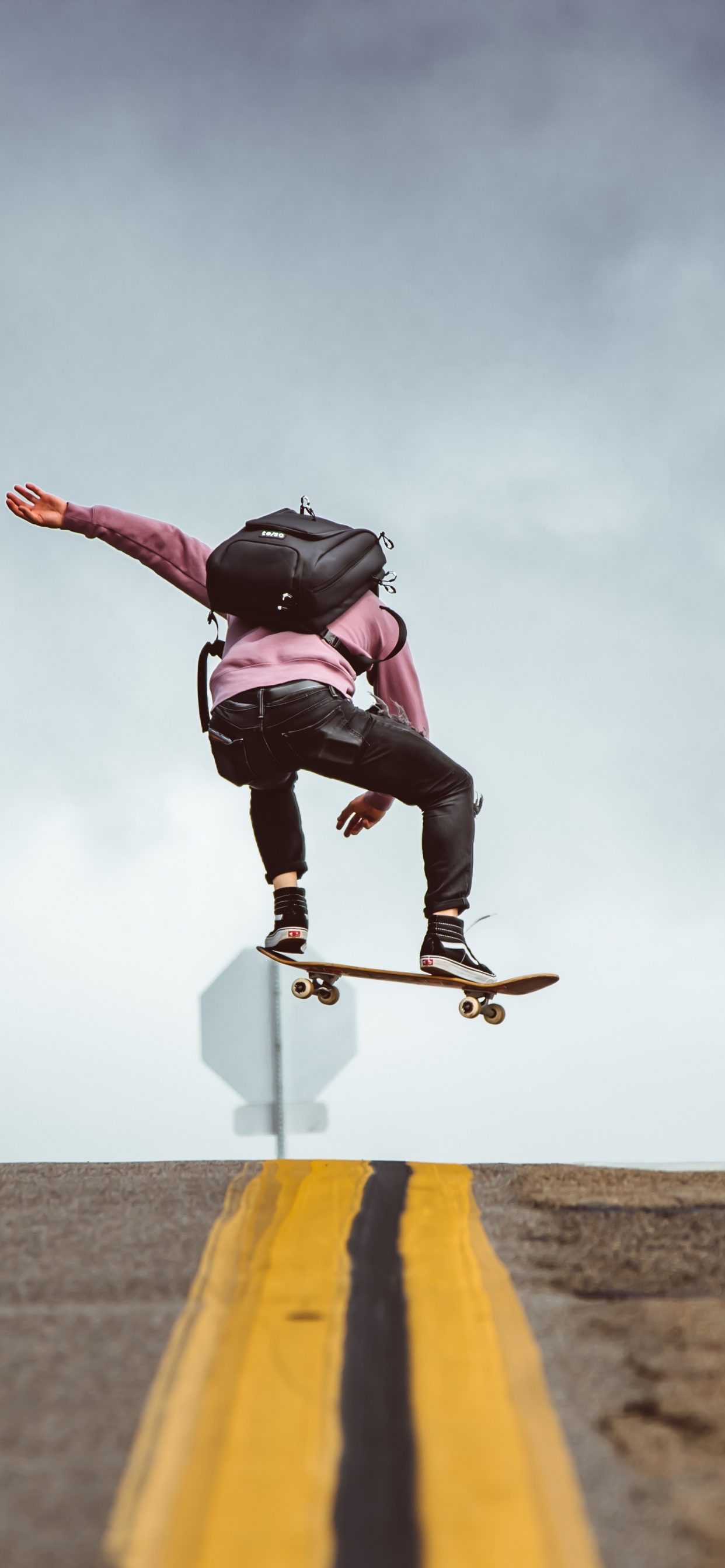 Mann in Schwarzer Jacke Und Schwarzer Hose Macht Stunts Auf Grauer Asphaltstraße Unter Blau Und. Wallpaper in 1242x2688 Resolution