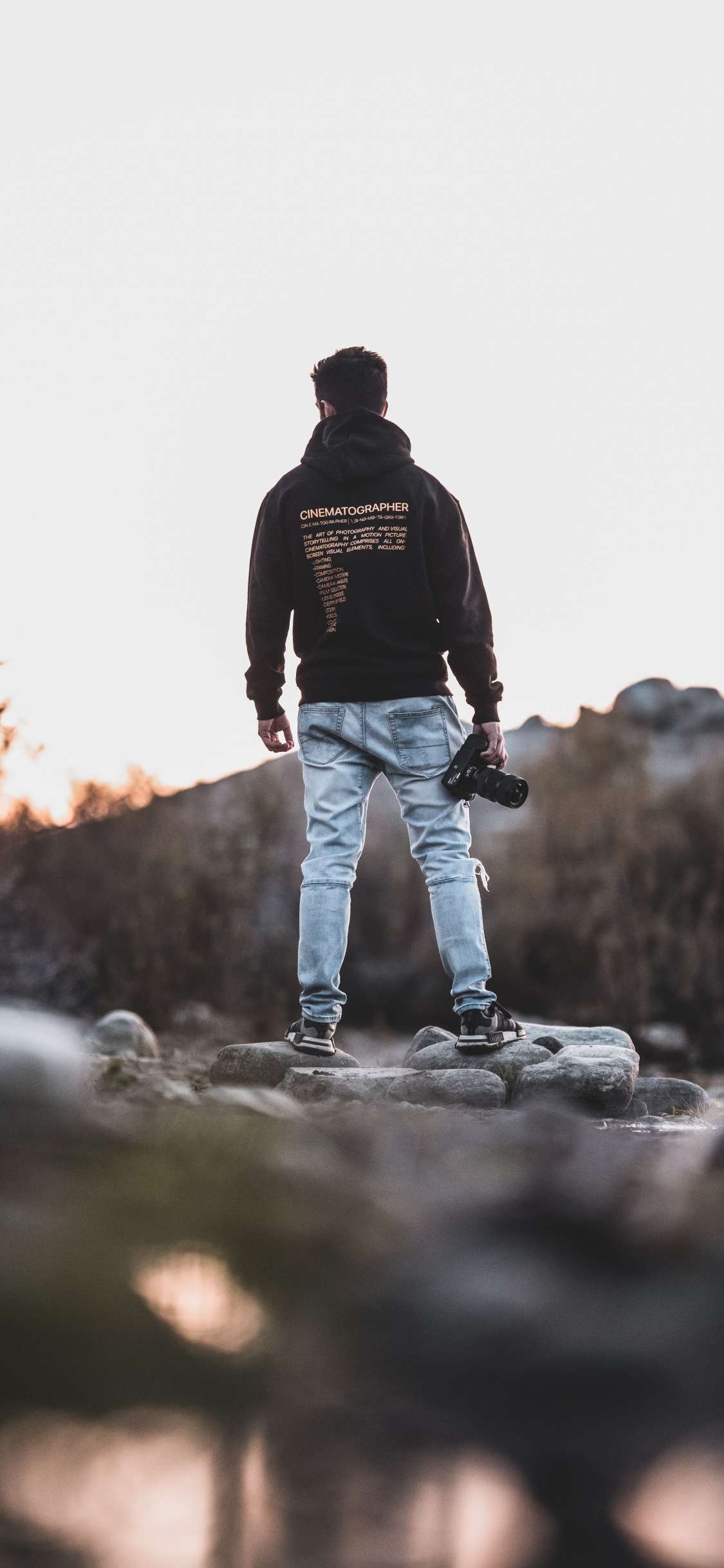 Hombre de Chaqueta Negra y Pantalones Vaqueros Azules Caminando Por la Carretera Rocosa Durante el Día. Wallpaper in 1125x2436 Resolution