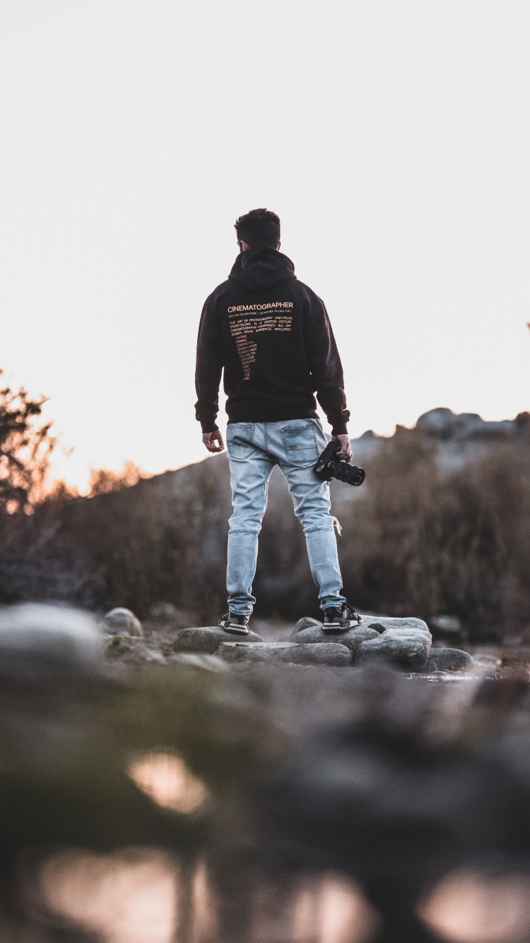 Man in Black Jacket and Blue Denim Jeans Walking on Rocky Road During Daytime. Wallpaper in 750x1334 Resolution