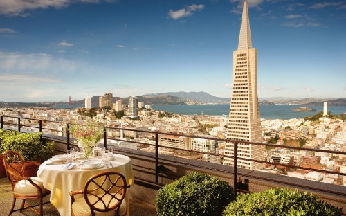 Image white and brown wooden table and chairs near green trees and city buildings during daytime