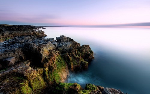 Image brown and green rock formation beside body of water