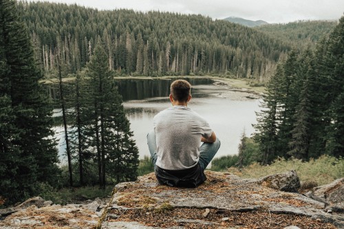 Image man in gray sweater sitting on brown rock near lake during daytime